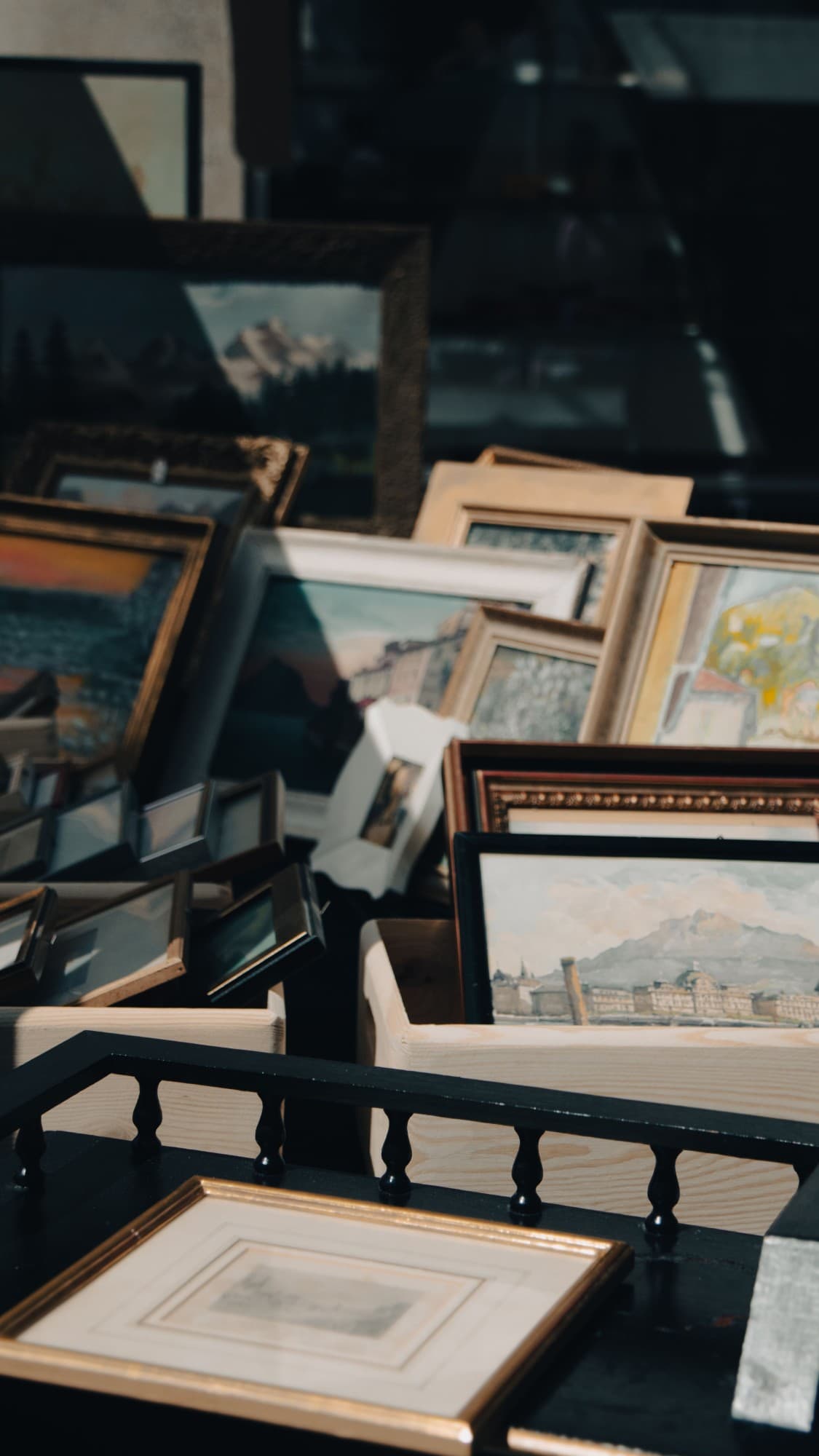 Framed paintings and prints stacked in wooden crates, lit by the sun